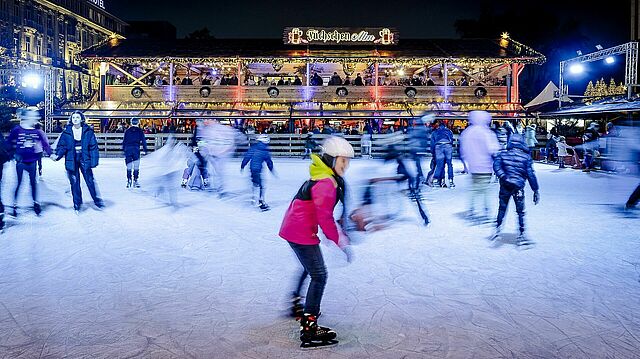 Kinder und Erwachsene fahren über die Eisfläche, mit der Alm im Hintergrund.