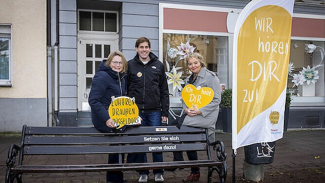 Die Inititatoren des Projekts sitzen auf einer Bank und halten ein Schild hoch.