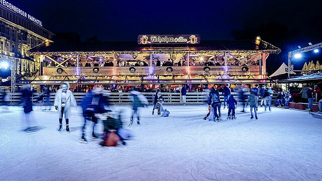 Menschen fahren Schlittschuh auf dem Eis. Im Hintergrund sieht man die Alm Terasse.